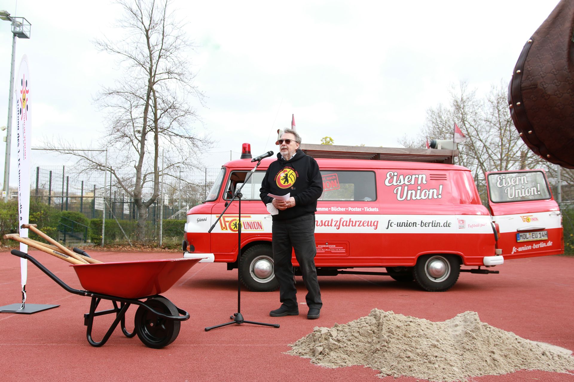 Mann mit Mikrofon vor einem roten Lieferwagen und einer Schubkarre auf einem Sportplatz, umgeben von Sand.