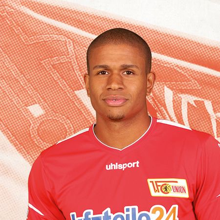 Ein Fußballspieler in rotem Trikot mit dem Logo von Union Berlin, vor einem orangefarbenen Hintergrund.