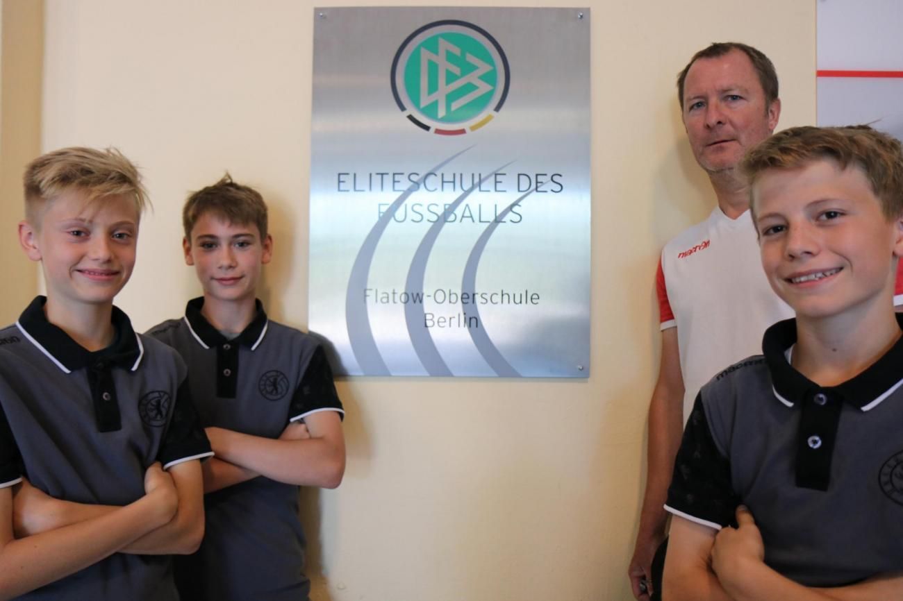 Drei Jungen in Sportkleidung und ein erwachsener Trainer stehen neben einem Schild der Eliteschule des Fußballs in Berlin.