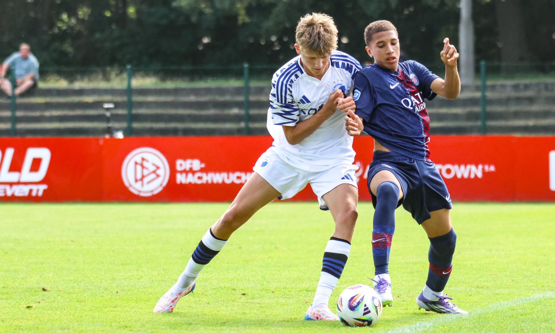 Zwei Fußballspieler in Aktion, während sie um den Ball kämpfen, auf einem grünen Spielfeld mit Werbebannern im Hintergrund.