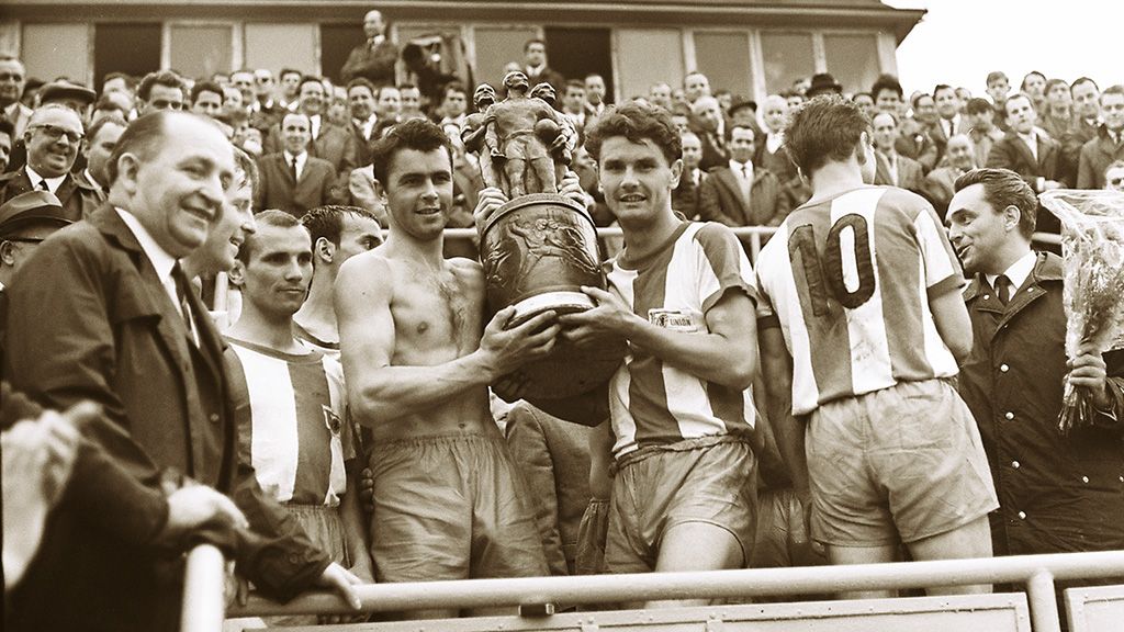 Two soccer players hold a trophy in front of cheering crowds while standing on a grandstand.