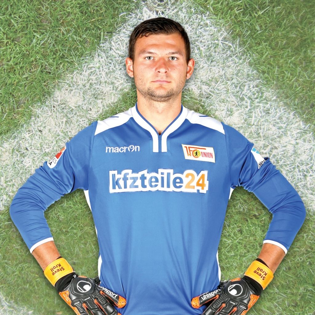 Ein Torwart steht im blauen Trikot mit Handschuhen auf einem Rasenfeld, Hände an den Seiten, Blick nach vorne.