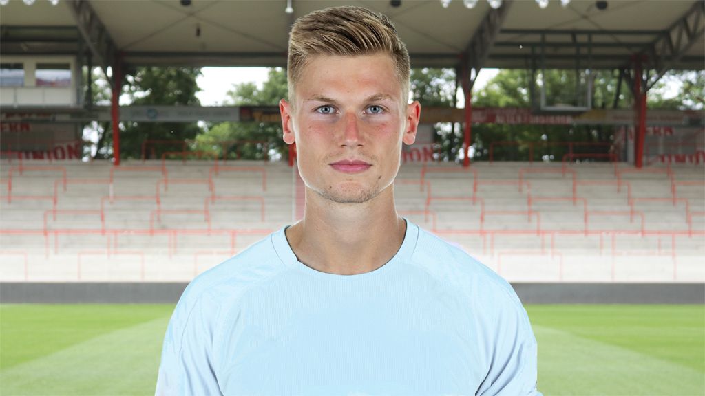 Junger Mann mit blonden Haaren in sportlicher Kleidung, steht vor einem Fußballstadion mit leeren Sitzplätzen im Hintergrund.