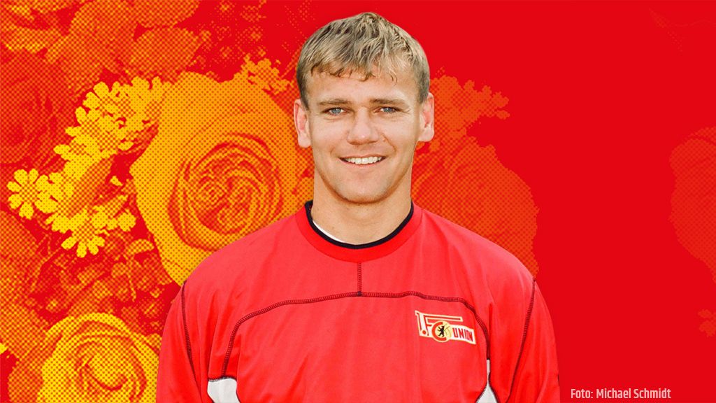 Ein lächelnder junger Mann in einem roten Sporttrikot steht vor einem bunten, floralem Hintergrund.