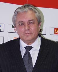 Ein Mann mit grauen Haaren in einem dunkel gestreiften Anzug und einer Krawatte, steht vor einem beschrifteten Hintergrund.