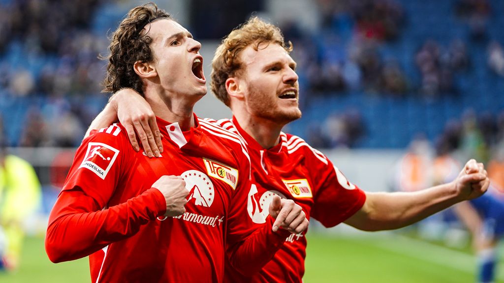 Two soccer players in red jerseys celebrate on the field, one with a raised fist and the other smiling.