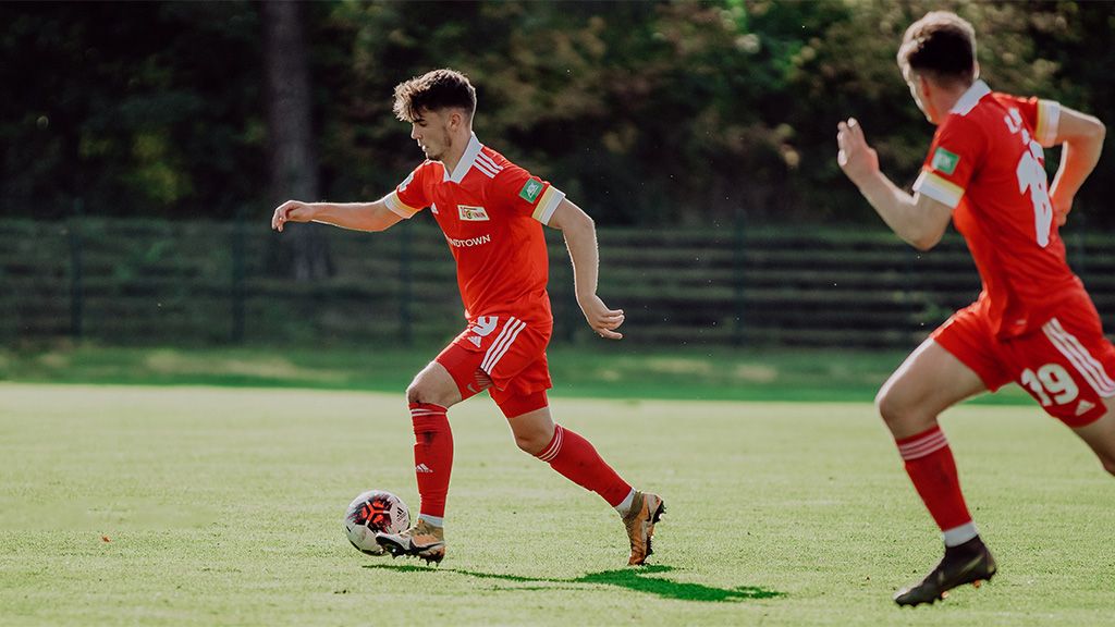 Ein Fußballspieler in rotem Trikot dribbelt den Ball auf einem Grünfeld, während ein Mitspieler im Hintergrund folgt.