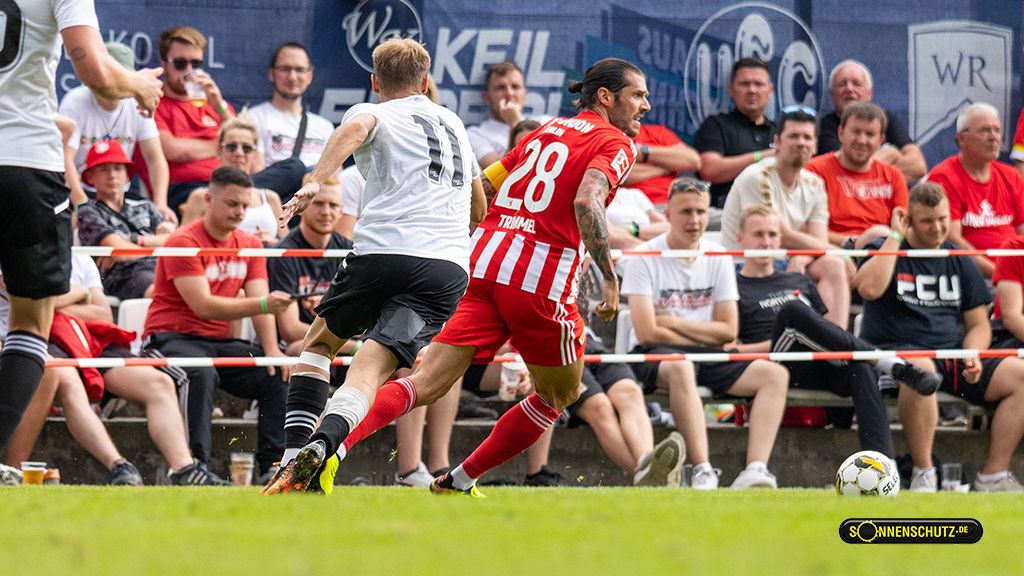 Ein Fußballspieler im roten Trikot verfolgt einen Mitspieler in Weiß, während Zuschauer auf den Tribünen zuschauen.