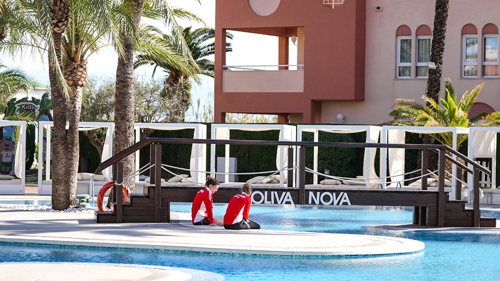 Zwei Personen in roten Shirts sitzen am Rand eines Pools mit Palmen und Liegen im Hintergrund.