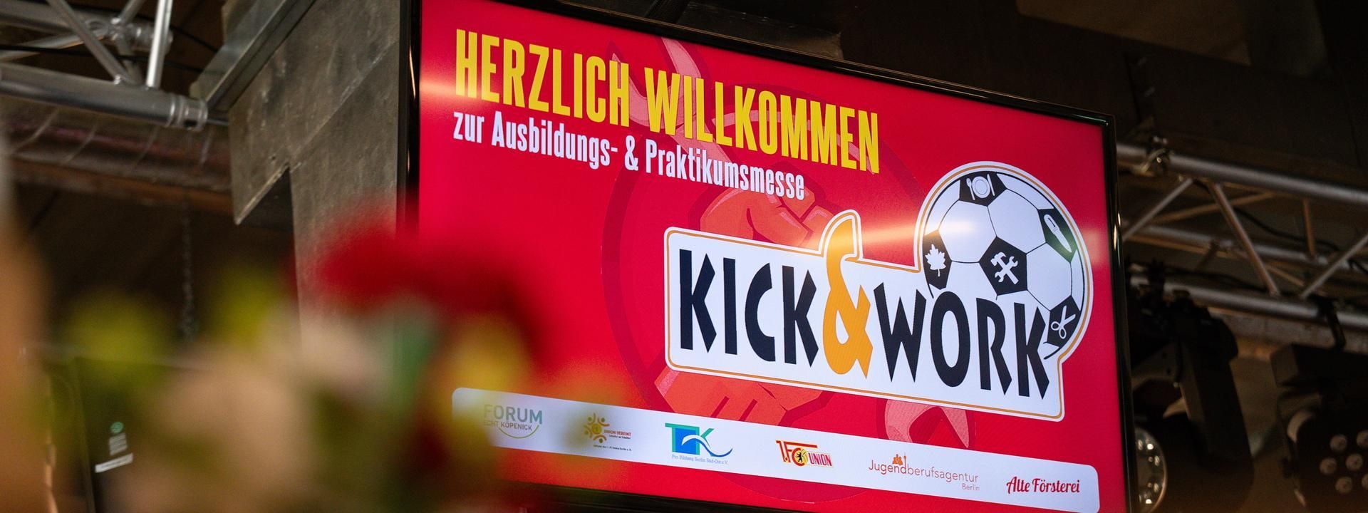 Ein Bildschirm zeigt den Schriftzug "Herzlich Willkommen zur Ausbildungs- & Praktikumsmesse Kick & Work" in einer festlichen Umgebung.