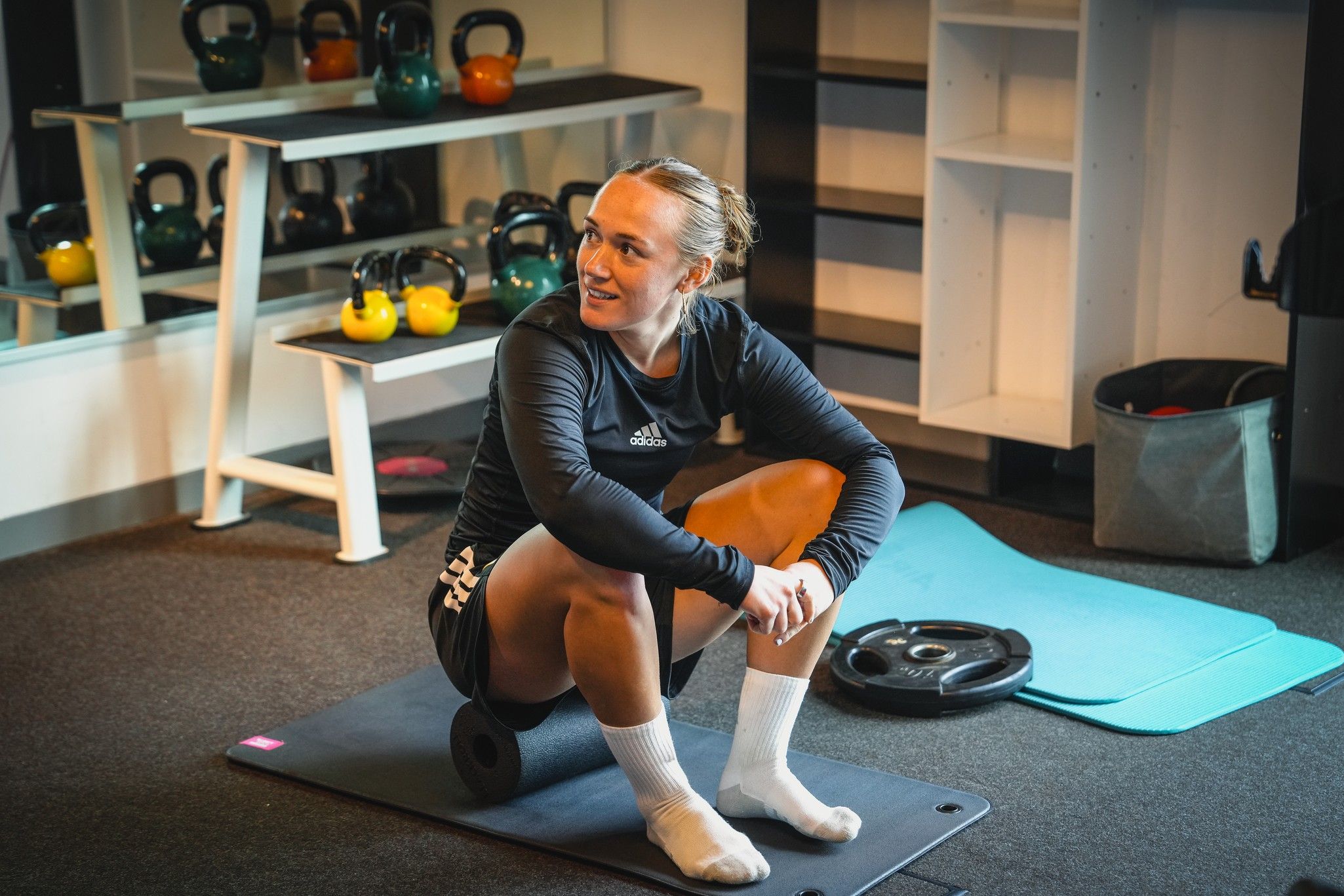 Eine Sportlerin sitzt auf einer Schaumstoffrolle in einem Fitnessraum, umgeben von Gewichten und Sportgeräten.