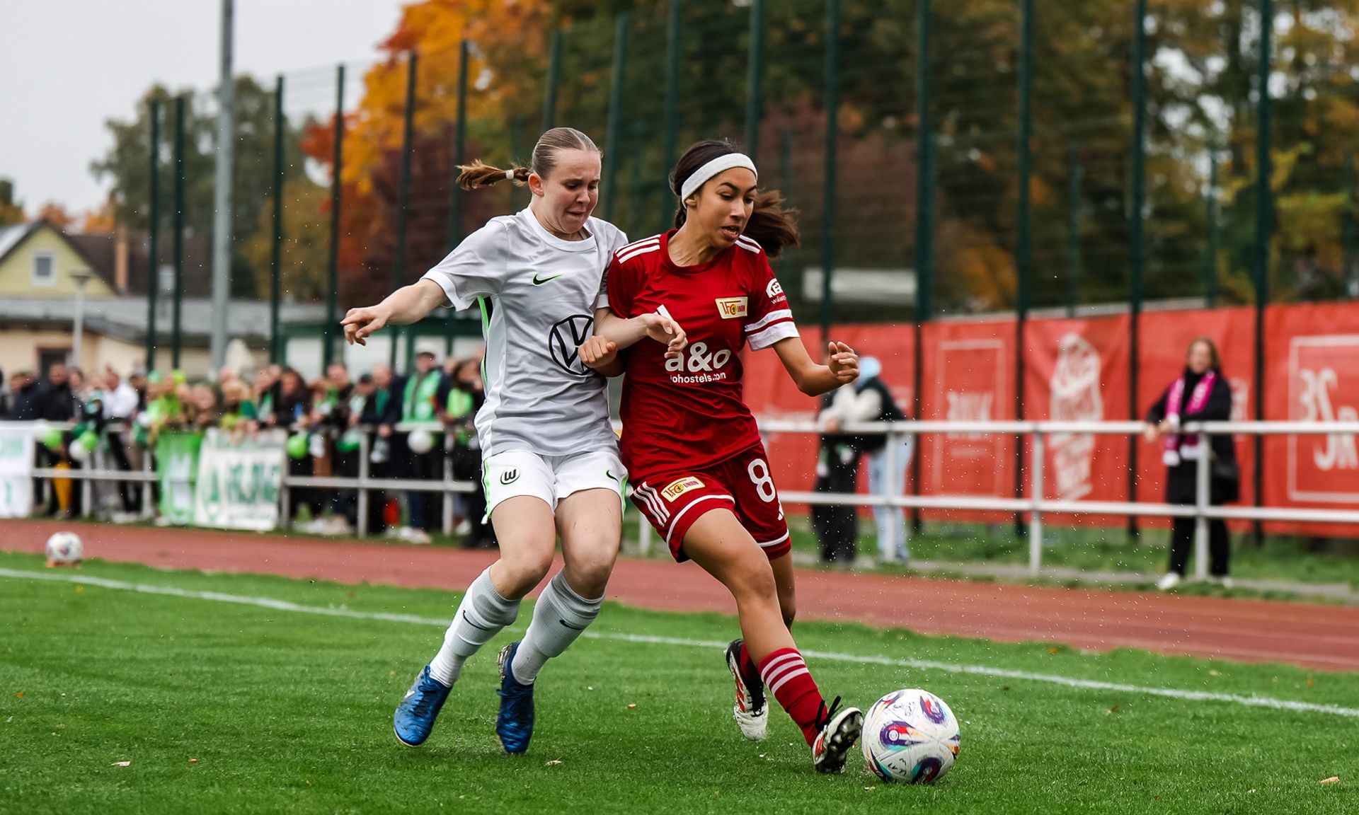 Eine Spielerin in rotem Trikot dribbelt mit dem Ball, während eine Gegnerin in weißem Trikot sie verfolgt. Herbstliche Umgebung im Hintergrund.