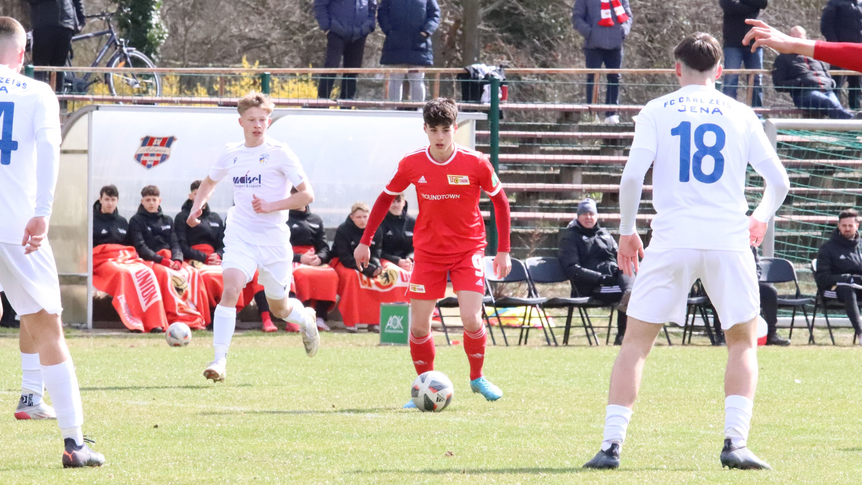 Ein Fußballspieler in rotem Trikot dribbelt den Ball, während zwei Spieler in weißen Trikots verteidigen. Zuschauer im Hintergrund.