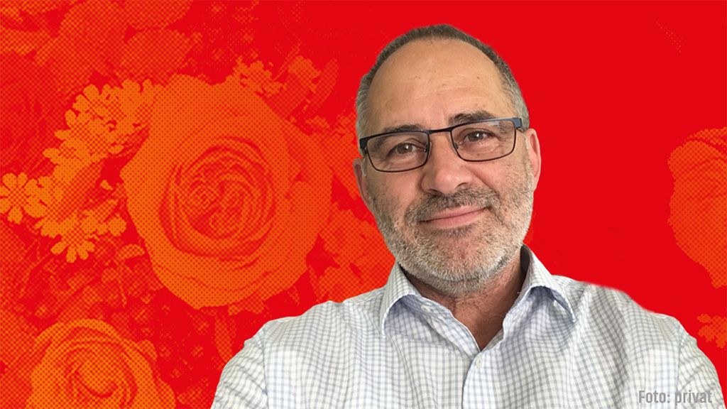 Mann mit Brille und Bart, lächelnd vor einem roten Hintergrund mit Blumenmuster.