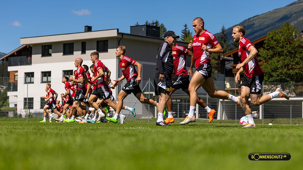 Fußballmannschaft in roten Trikots beim Training auf einem grünen Feld, umgeben von Bergen und einem Gebäude im Hintergrund.