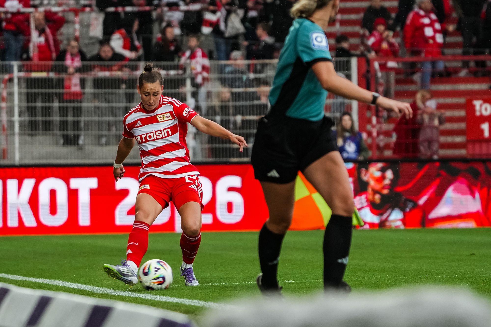 Eine Fußballspielerin in rot-weiß gestreiftem Trikot führt einen Eckball aus, während eine Schiedsrichterin in der Nähe steht.