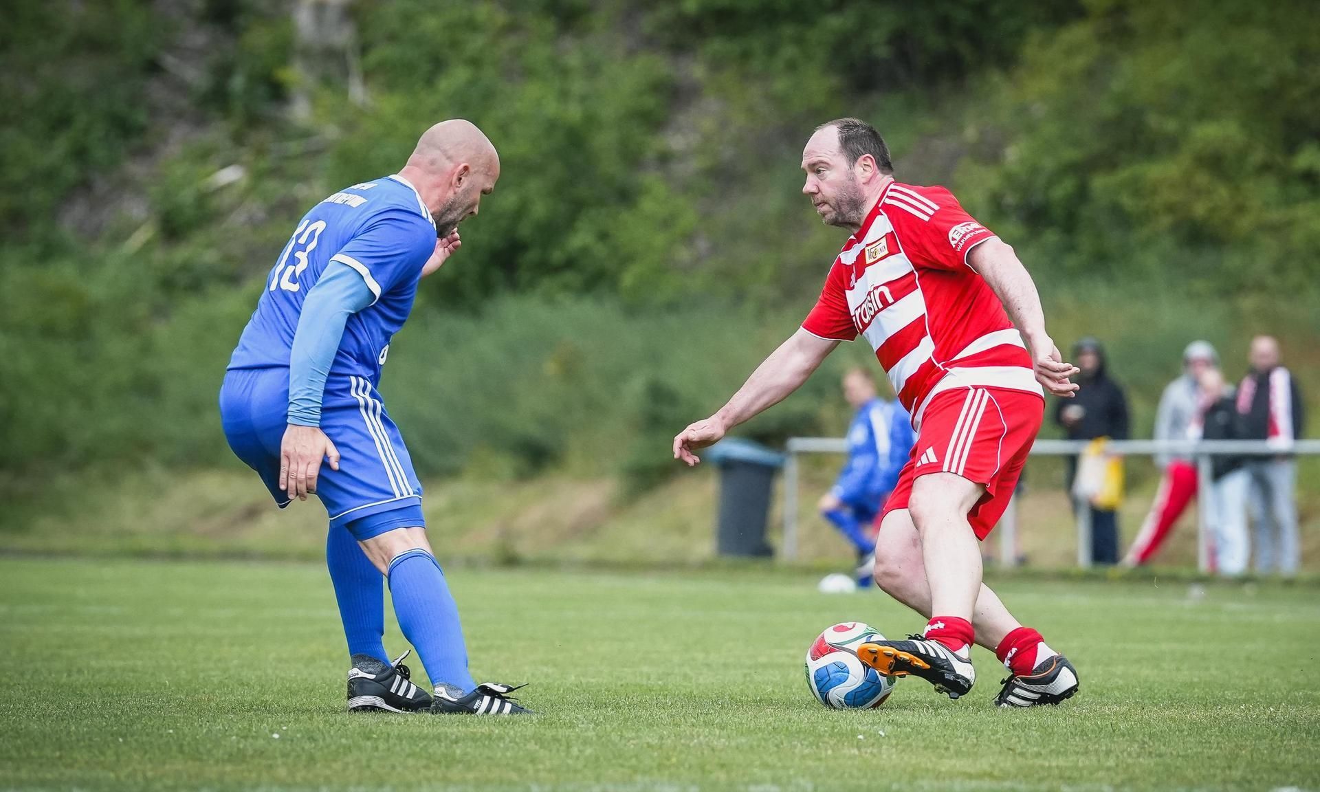 Zwei Fußballspieler in einem Spiel: einer in blauer, der andere in roter Uniform, auf einem grünen Rasenfeld.