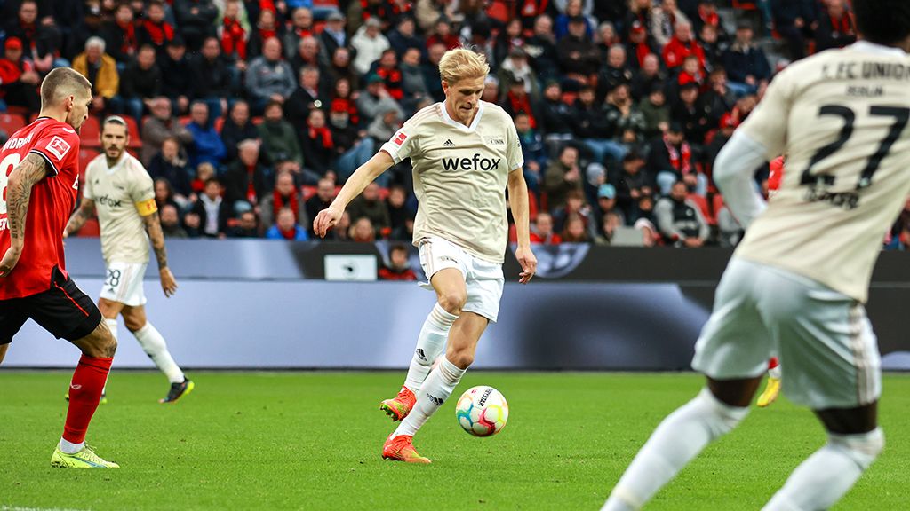 A player in a beige jersey shoots the ball during a soccer game, with spectators visible in the background.
