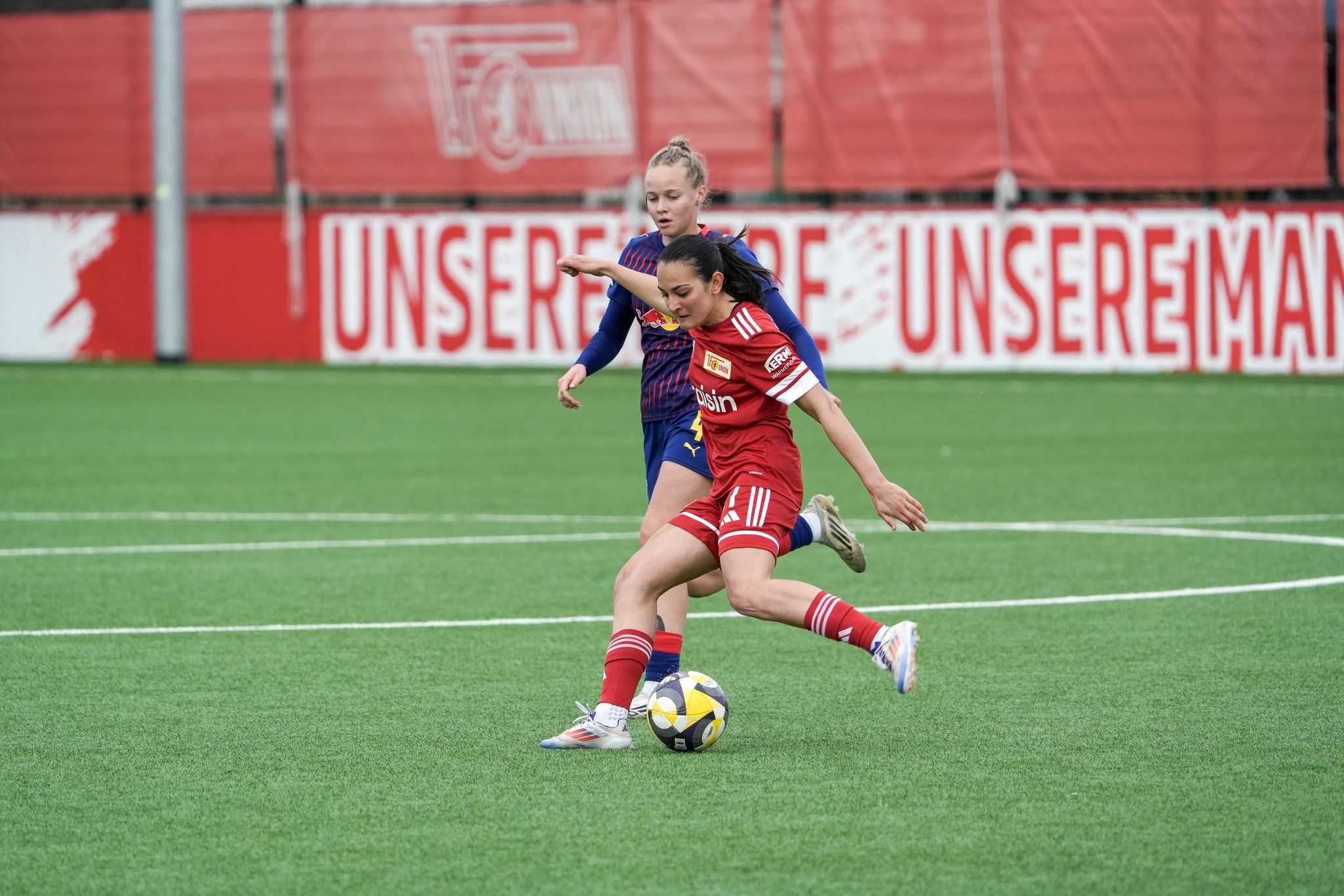 Eine Spielerin in rotem Trikot tritt den Ball, während eine Gegnerin in blauem Trikot sie verfolgt.