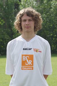 Junger Mann mit lockigem Haar, der ein weißes Fußballtrikot trägt, steht auf einem Sportplatz vor grünen Bäumen.