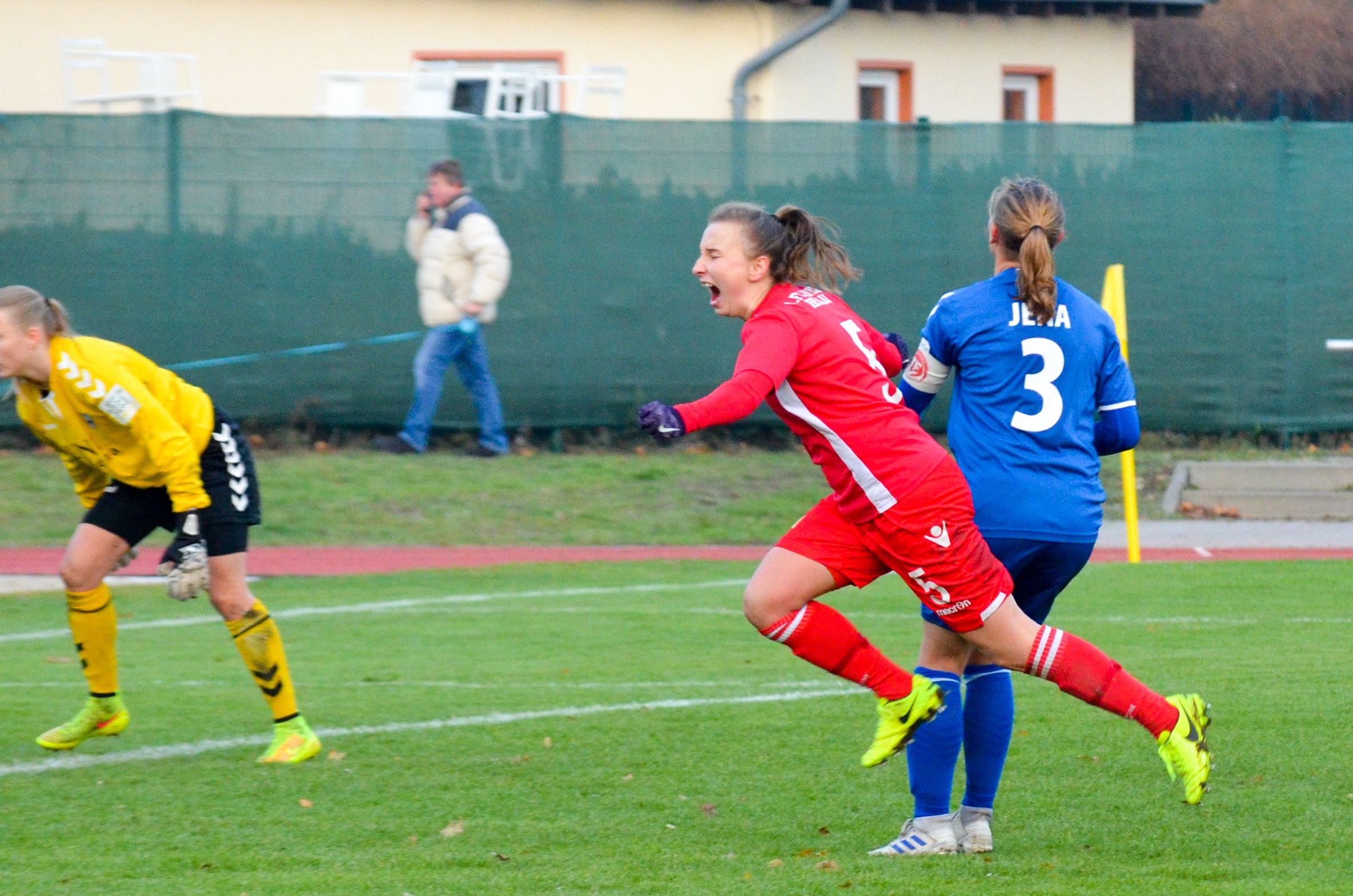 Eine Spielerin in rotem Trikot läuft fröhlich. Sie ist auf dem Platz. Die Torhüterin trägt Gelb. Eine andere Spielerin trägt Blau. Sie stehen im Hintergrund.