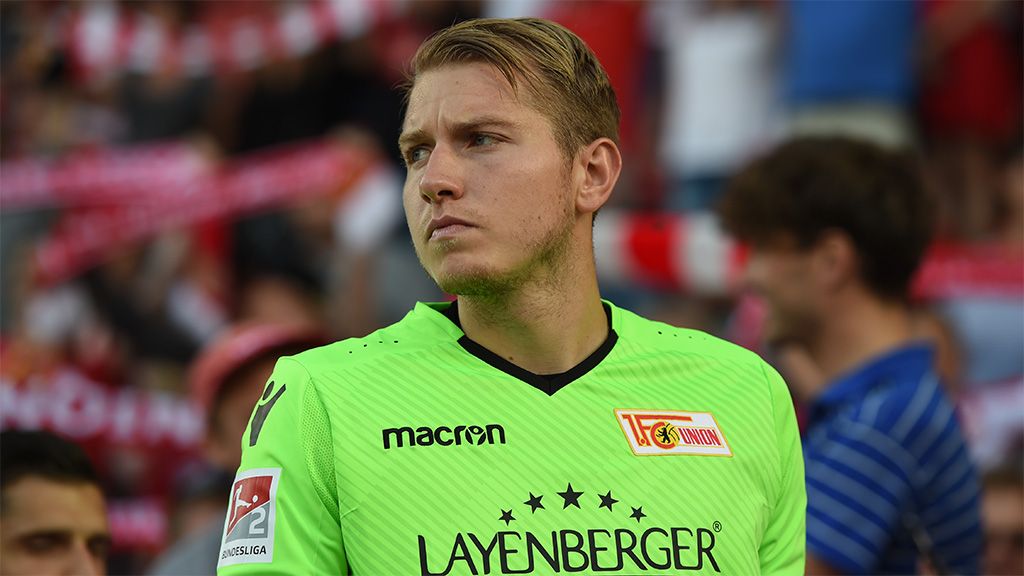 Ein Torwart in einem hellgrünen Trikot steht auf dem Platz und schaut ernst in die Ferne. Im Hintergrund sind Fans und Stadion zu sehen.