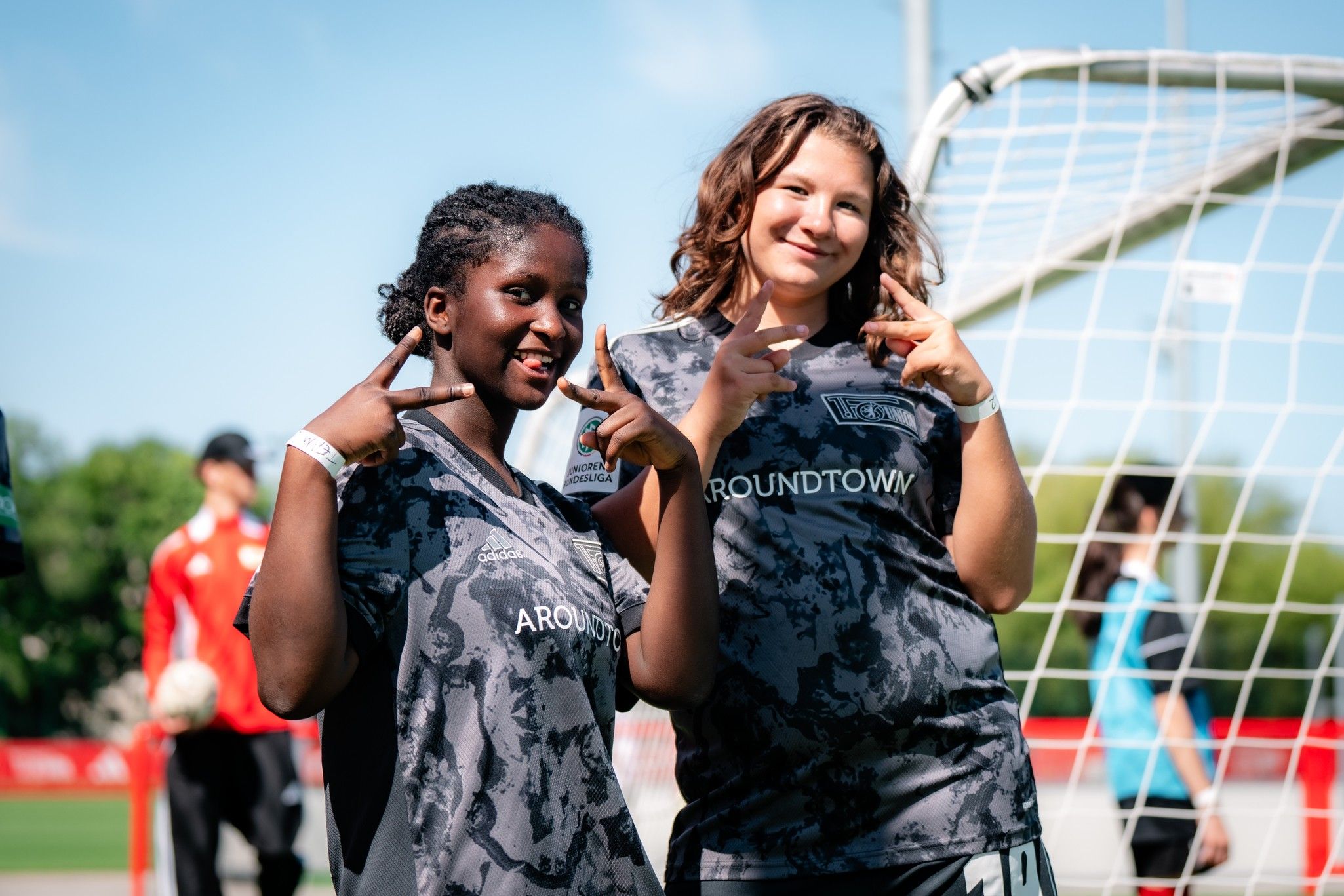 Zwei Mädchen in Fußballtrikots posieren lächelnd vor einem Tor auf einem Sportplatz.