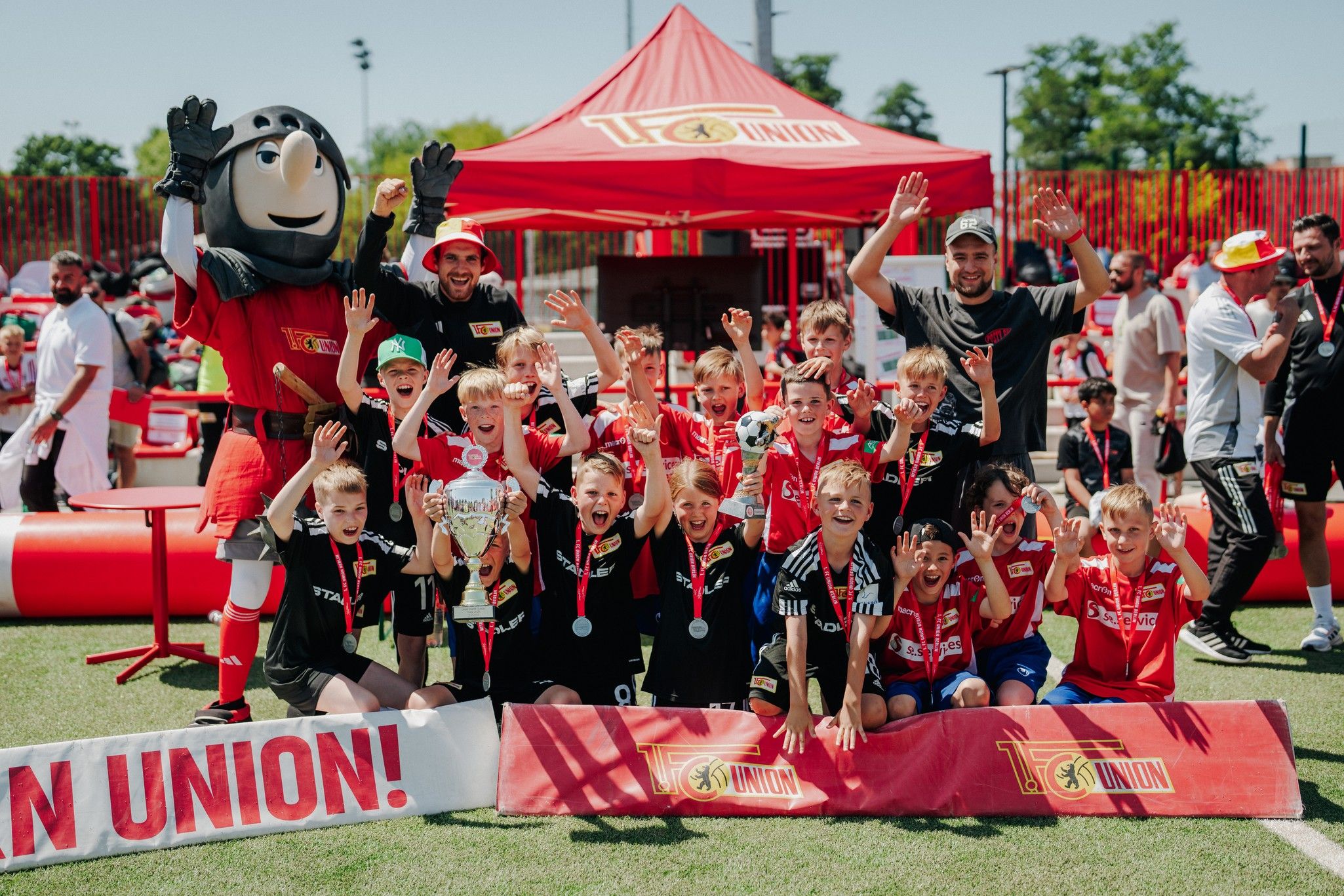 Eine Gruppe junger Fußballspieler jubelt auf einem Platz. Im Hintergrund steht ein Zelt mit dem Logo des Vereins und ein Maskottchen.