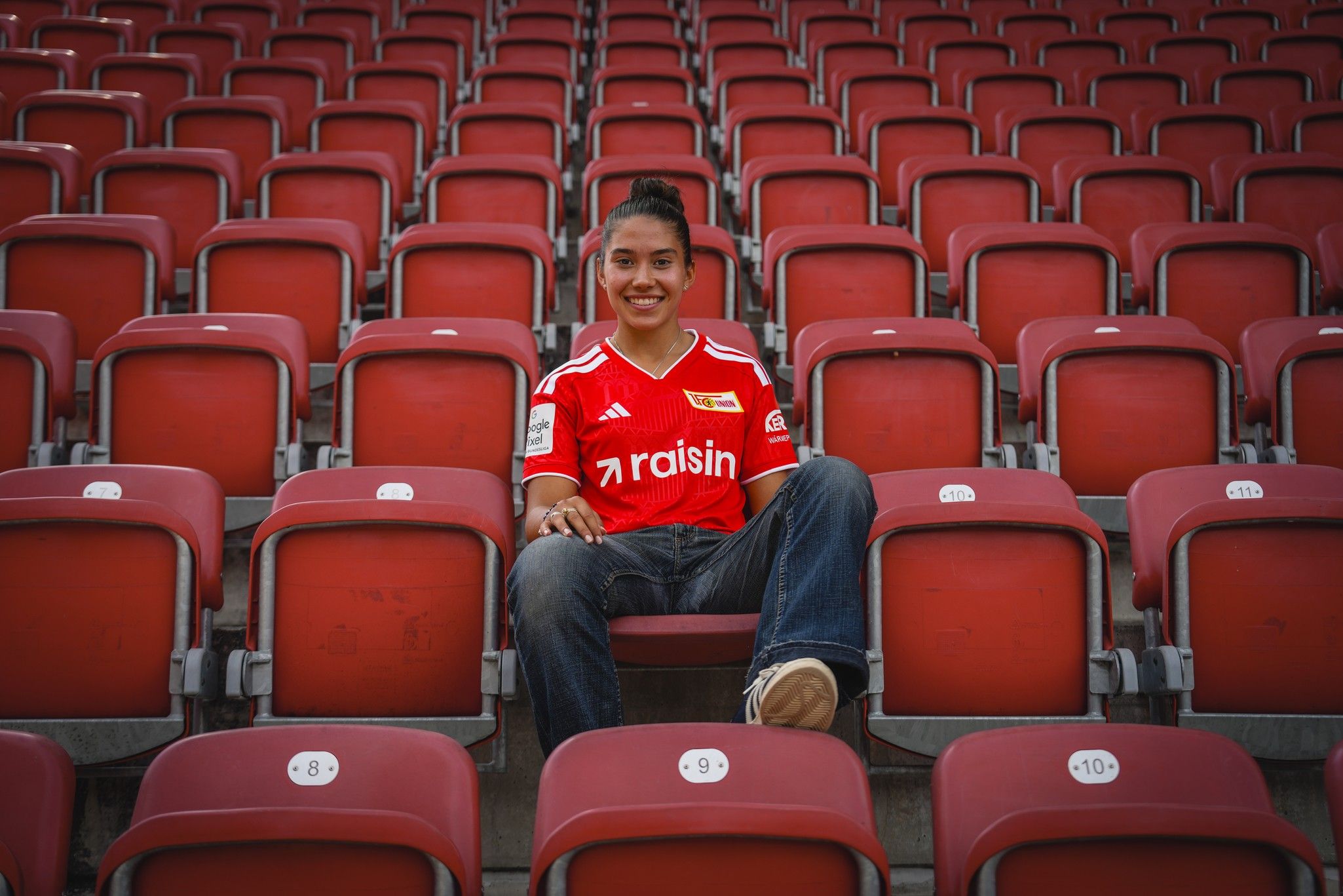 Ein junger Mensch sitzt entspannt in einem fast leeren Stadion, trägt ein rotes Trikot und lächelt in die Kamera.