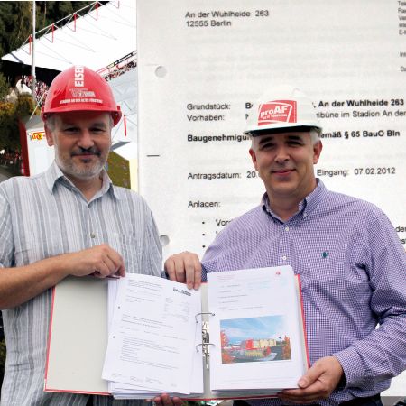 Zwei Männer in Schutzhelmen halten einen Ordner mit Bauunterlagen vor einem Bauprojekt in Berlin.