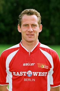 Ein männlicher Fußballspieler trägt ein rotes Trikot mit weißen Details und lächelt in die Kamera.