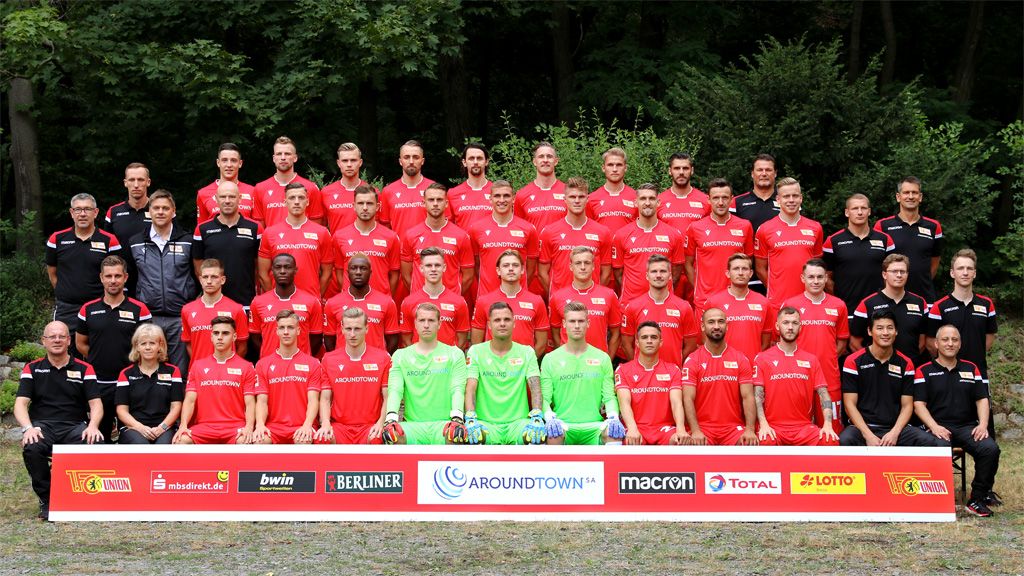 Gruppenfoto einer Fußballmannschaft in roten Trikots, umgeben von Trainern und Betreuern, vor einer grünen Waldkulisse.