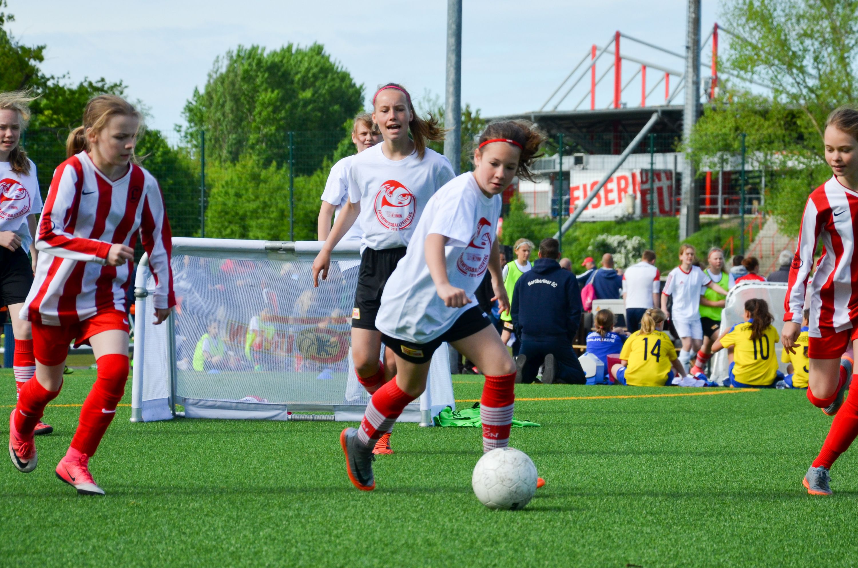 Mädchen spielen Fußball auf einem Kunstrasenplatz, einige tragen rote und weiße Trikots, andere gelbe Trikots im Hintergrund.