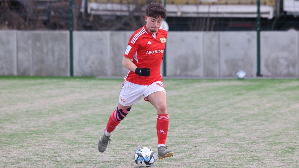 Ein junger Fußballspieler in rotem Trikot dribbelt einen Ball auf einem grünen Rasenfeld.