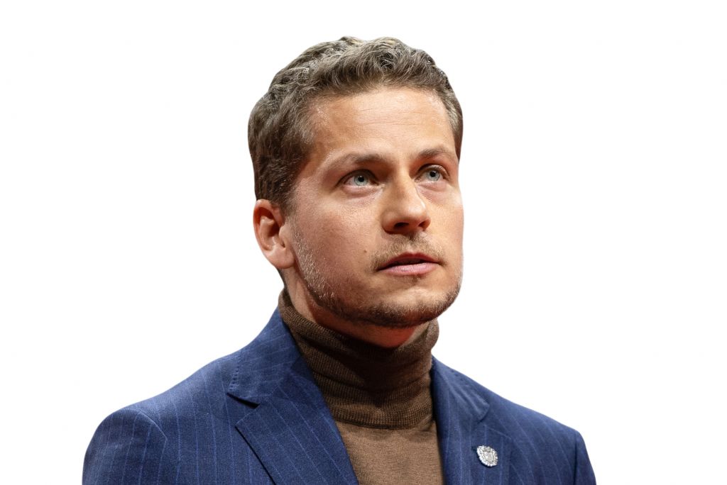 Ein Mann mit kurzen, blonden Haaren und einem Bart trägt einen grauen Anzug und einen braunen Rollkragenpullover, vor einem weißen Hintergrund.