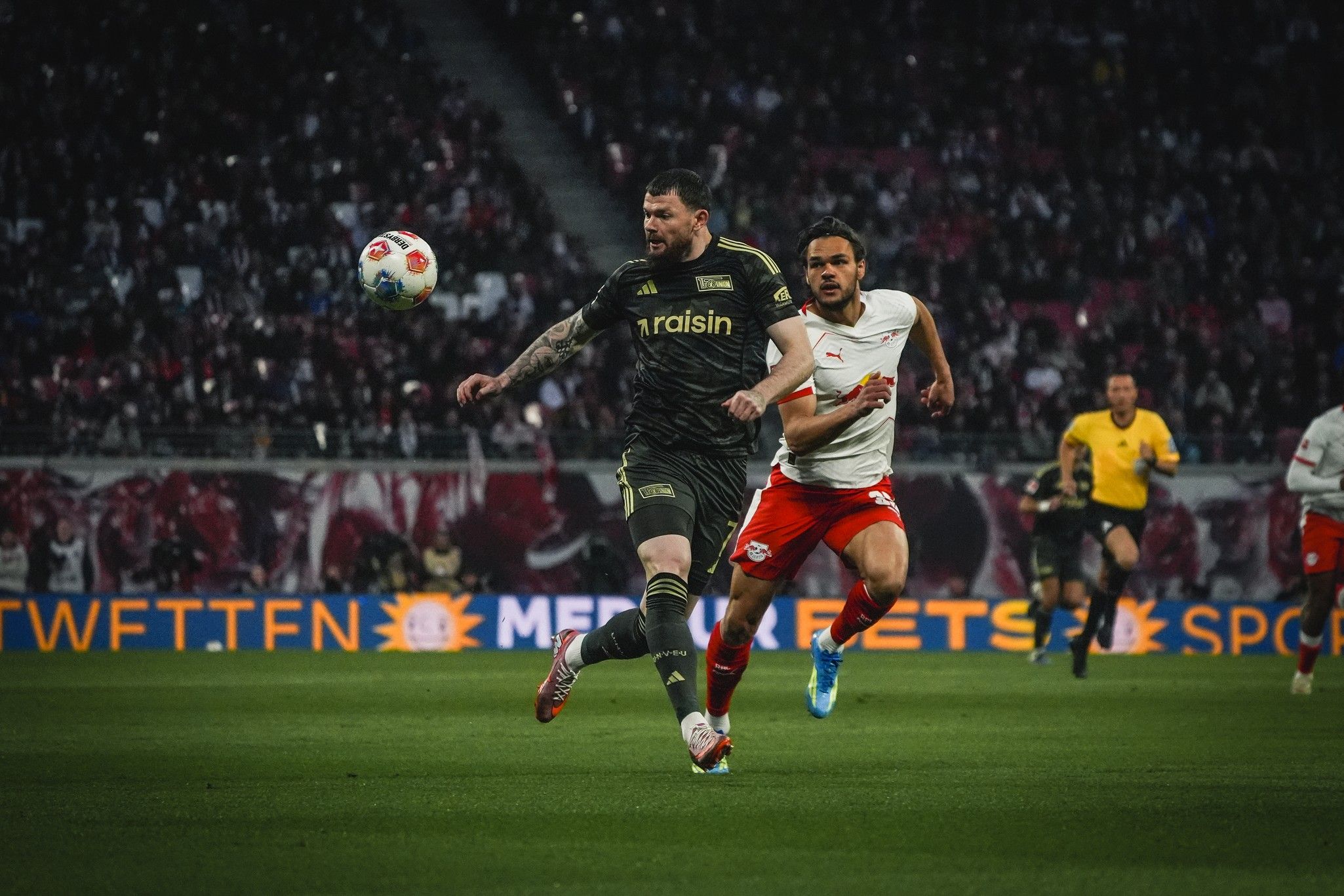 Ein Fußballspieler in dunkler Uniform spielt den Ball, während ein Spieler in roter Uniform ihn verfolgt. Zuschauer im Hintergrund.
