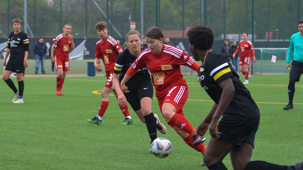 Mädchenfußballspiel auf einem Kunstrasenplatz, Spielerinnen in roten und schwarzen Trikots dribbeln den Ball.