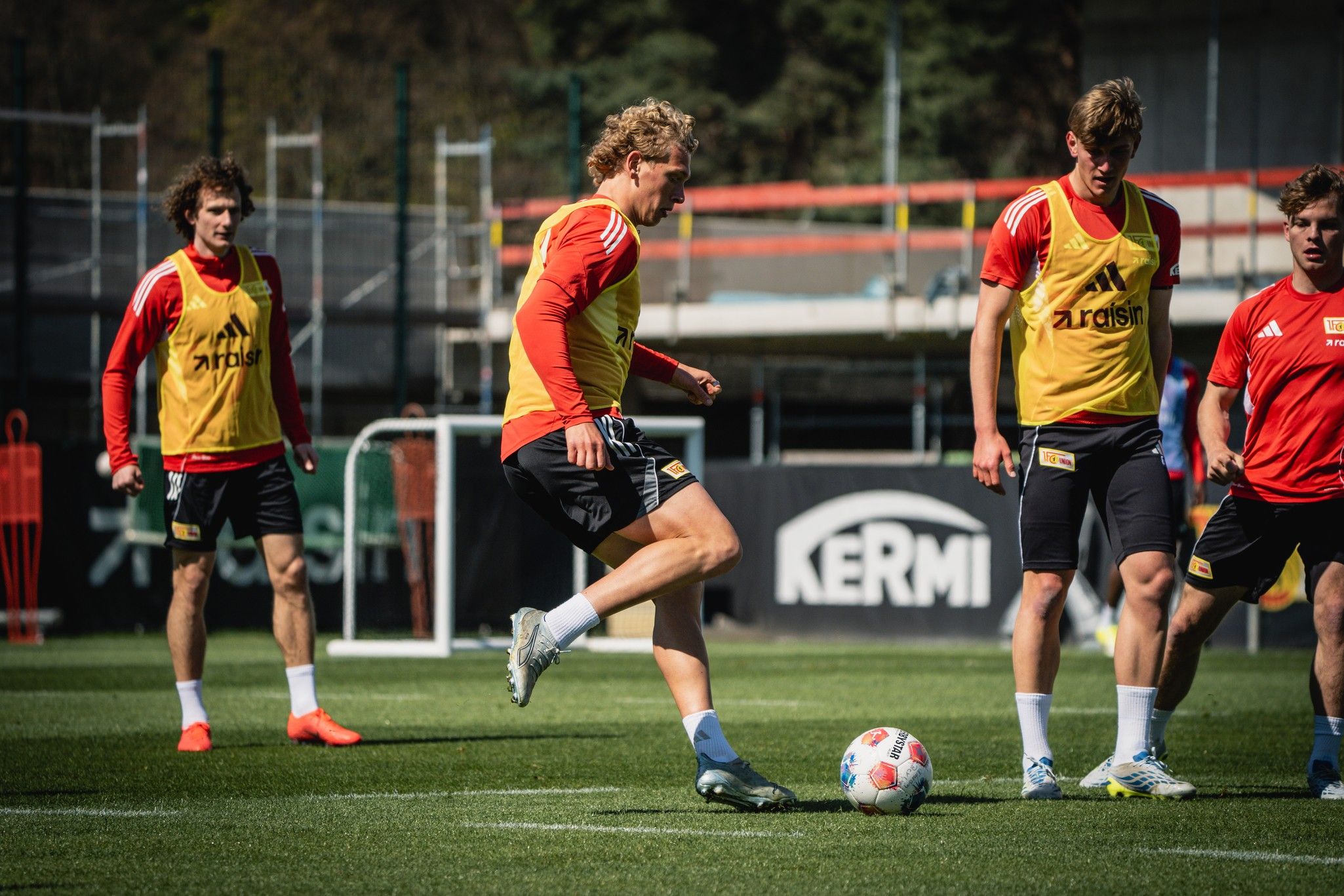 Fußballspieler trainieren auf dem Platz, einige in gelben, andere in roten Trikots, während sie einen Ball spielen.