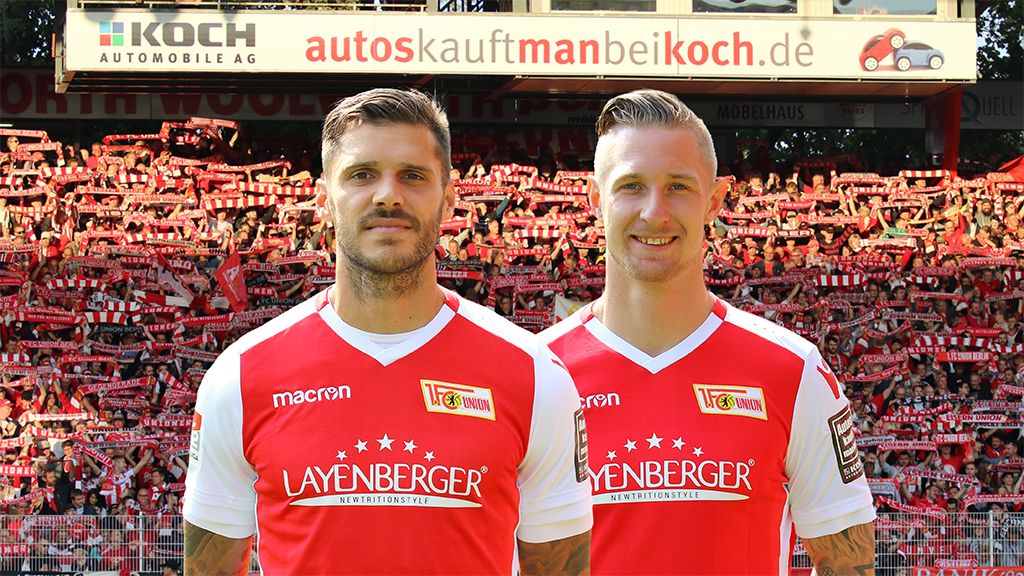 Zwei Fußballspieler in roten Trikots stehen vor einer Menge von Fans mit Schals im Hintergrund, die ein Stadion unterstützen.