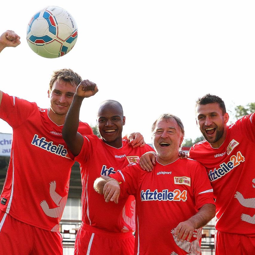 Vier Personen in roten Fußballtrikots stehen lächelnd zusammen und heben ihre Arme in die Luft. Ein Fußball ist in der Mitte der Gruppe.
