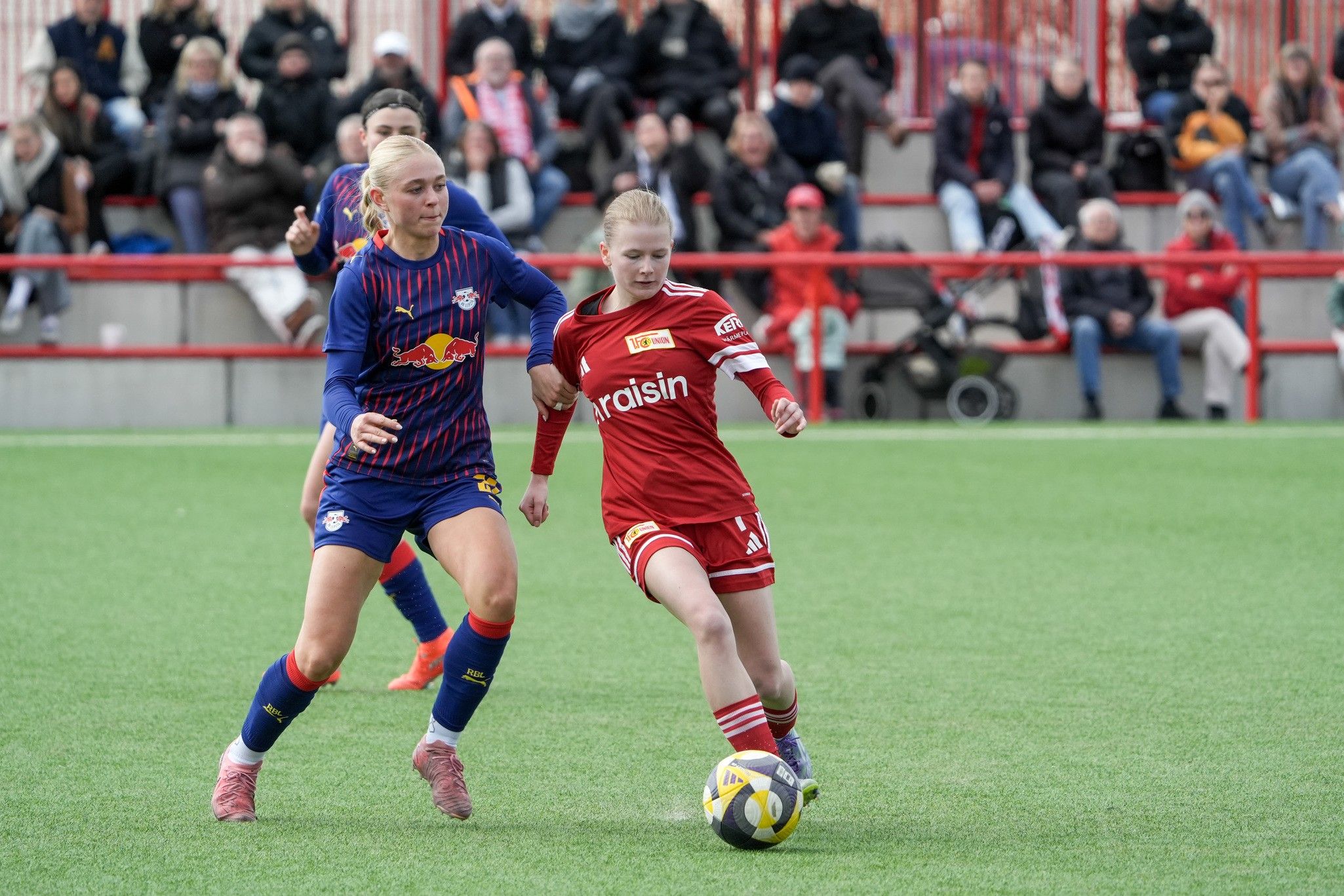 Zwei Fußballspielerinnen kämpfen um den Ball auf einem Rasenplatz, während Zuschauer im Hintergrund zuschauen.