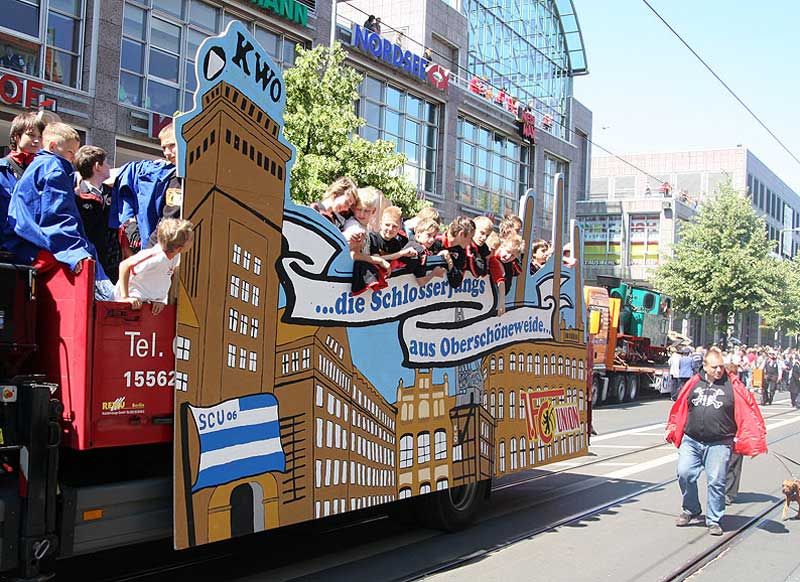 Ein bunt dekorierter Festwagen mit Kindern, der durch eine Stadtstraße fährt, umgeben von Zuschauern und Gebäuden.