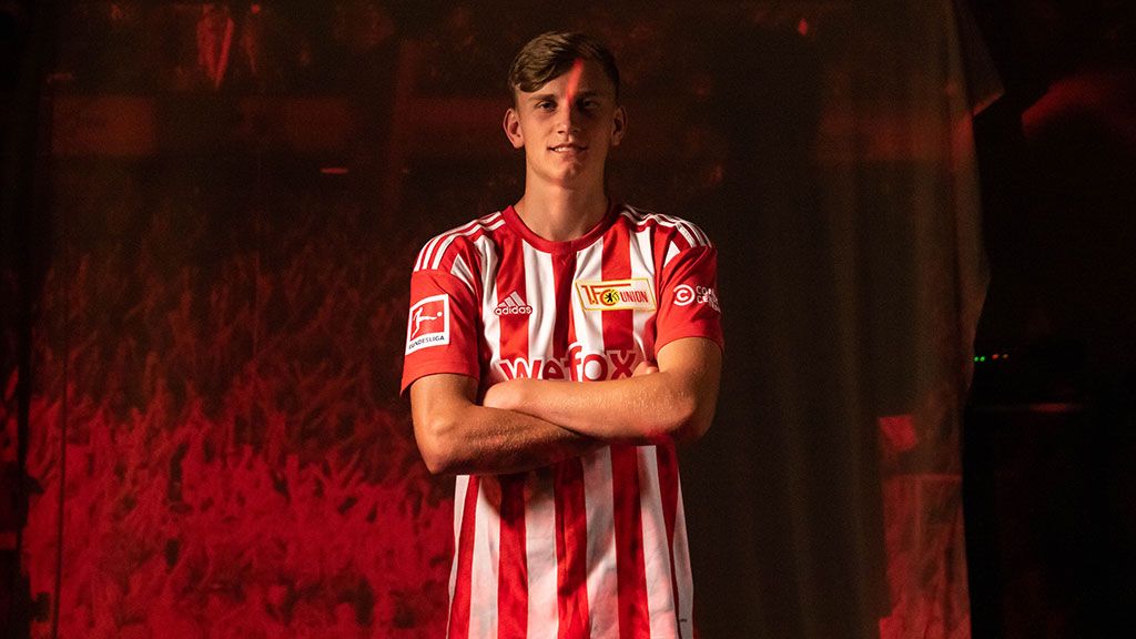 Ein junger Fußballspieler steht vor einem dunklen Hintergrund. Er trägt ein rot-weißes Trikot. Seine Arme sind gekreuzt.