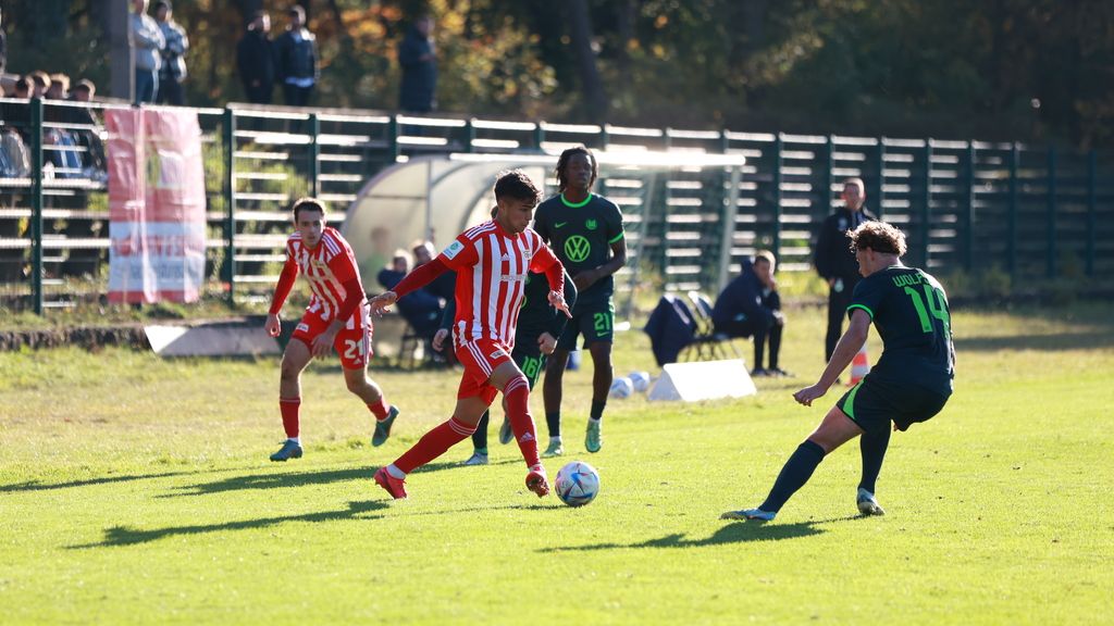 Ein Fußballspieler im rot-weißen Trikot dribbelt den Ball, während zwei Gegenspieler ihn verfolgen. Zuschauer stehen im Hintergrund.