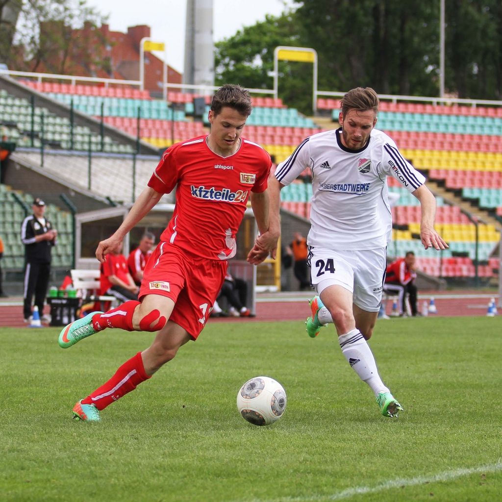 Zwei Fußballspieler im Wettkampf um den Ball auf einem Stadionrasen, mit Zuschauern im Hintergrund.