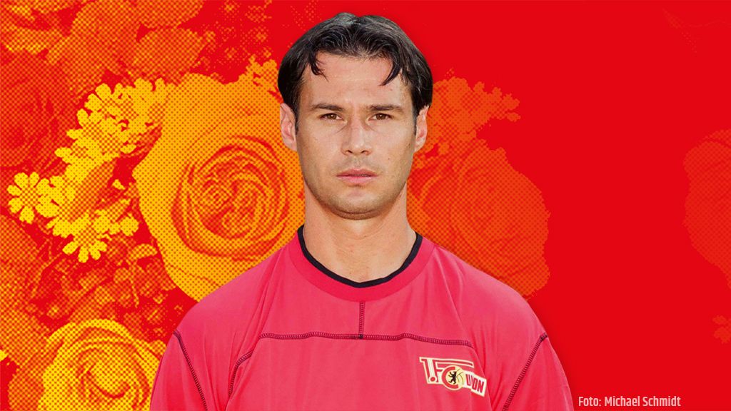 Ein Mann in rotem Trikot mit kurzem, dunklem Haar vor einem rot-blütenmusternden Hintergrund.