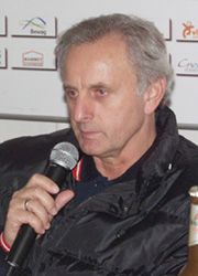 Mann mit grauen Haaren hält ein Mikrofon und trägt eine schwarze Jacke, vor einem Hintergrund mit Logos.