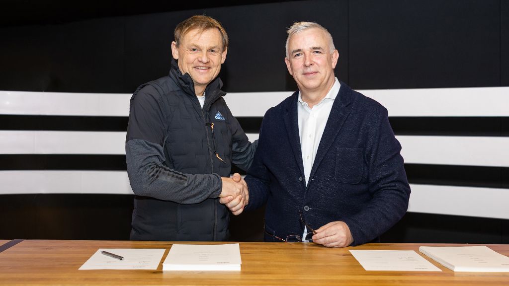 Two men shake hands in front of a table with documents and a black wall in the background.