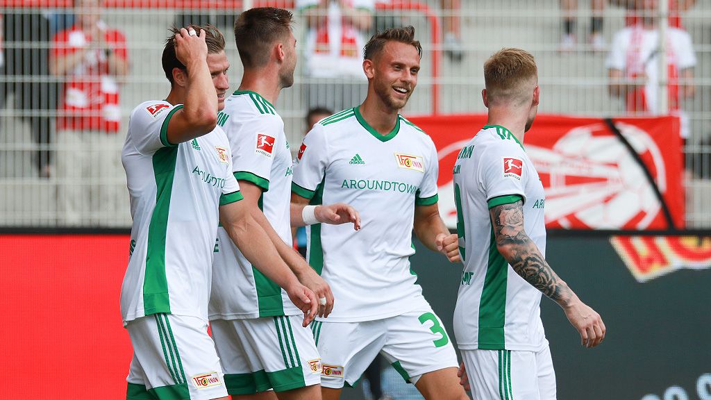 Four soccer players in white jerseys with green accents are smiling together on the field.