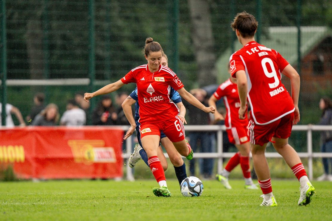 Zwei Spielerinnen in roten Trikots spielen Fußball auf einem Rasenplatz, während Zuschauer im Hintergrund beobachten.